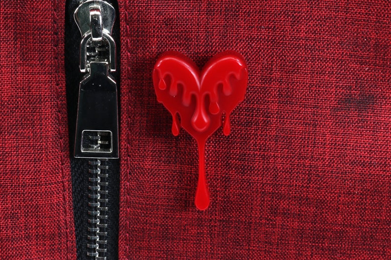 May include: A red heart-shaped pin with dripping red paint. The pin is attached to a red fabric background.