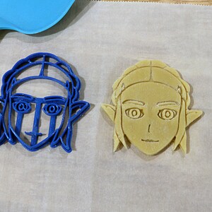 May include: Two cookie cutters, one blue and one beige, shaped like a person's face with elf ears and hair.