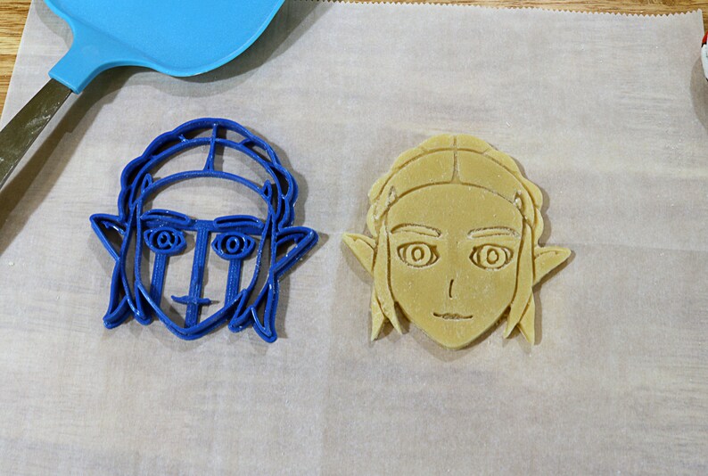 May include: Two cookie cutters, one blue and one beige, shaped like a person's face with elf ears and hair.