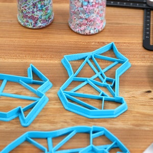 May include: Four blue plastic cookie cutters shaped like geometric designs. The cutters are on a wooden surface.