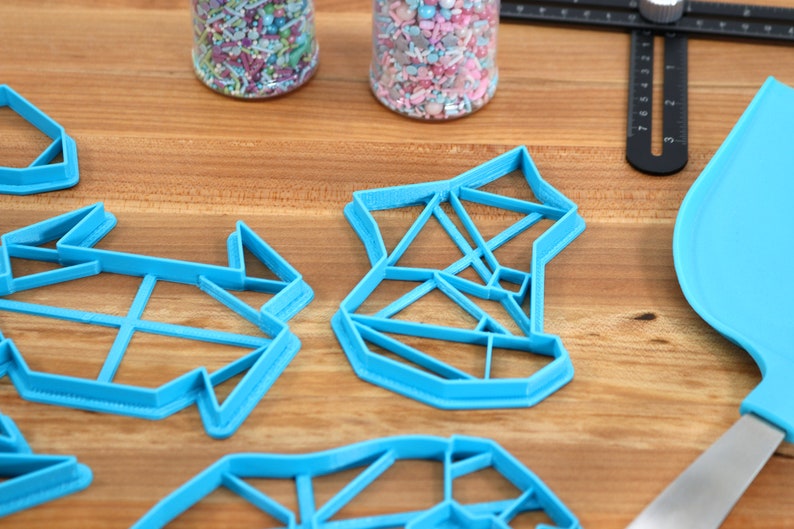 May include: Four blue plastic cookie cutters shaped like geometric designs. The cutters are on a wooden surface.