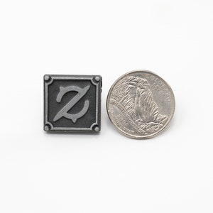 Op de afbeelding: Een zilverkleurige metalen pin met een gestileerde letter Z in een vierkant. De pin is ongeveer zo groot als een Amerikaanse kwart.