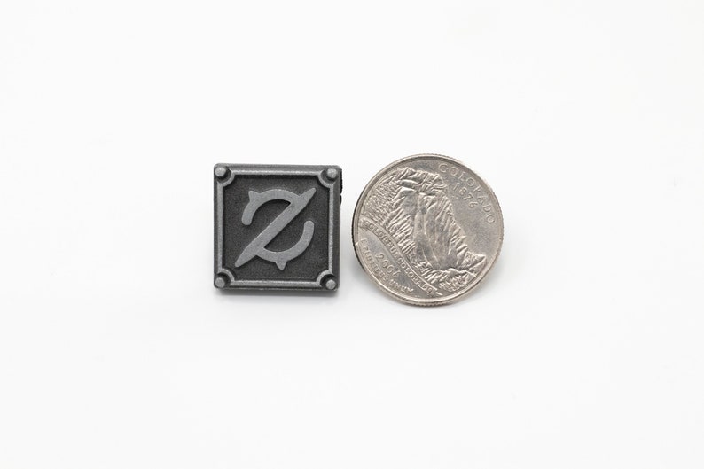 Op de afbeelding: Een zilverkleurige metalen pin met een gestileerde letter Z in een vierkant. De pin is ongeveer zo groot als een Amerikaanse kwart.