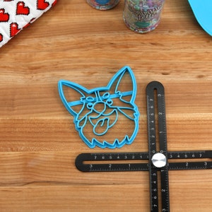 Pu&ograve; includere: Tagliapasta in plastica blu a forma di testa di corgi con la lingua che sporge. Il tagliapasta &egrave; su una superficie di legno con un righello nero.