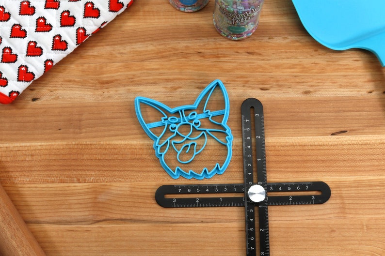 Pu&ograve; includere: Tagliapasta in plastica blu a forma di testa di corgi con la lingua che sporge. Il tagliapasta &egrave; su una superficie di legno con un righello nero.