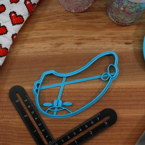 May include: A blue plastic cookie cutter shaped like a cartoon face with a frown. The cookie cutter is on a wooden surface.