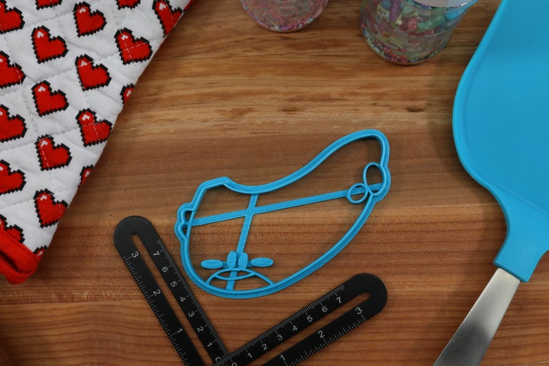 May include: A blue plastic cookie cutter shaped like a cartoon face with a frown. The cookie cutter is on a wooden surface.