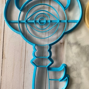 Op de afbeelding: Een blauwe plastic koekjesvormer in de vorm van een sleutel met een cirkelvormig oogontwerp. Het oog heeft concentrische cirkels en een kleine cirkel in het midden.