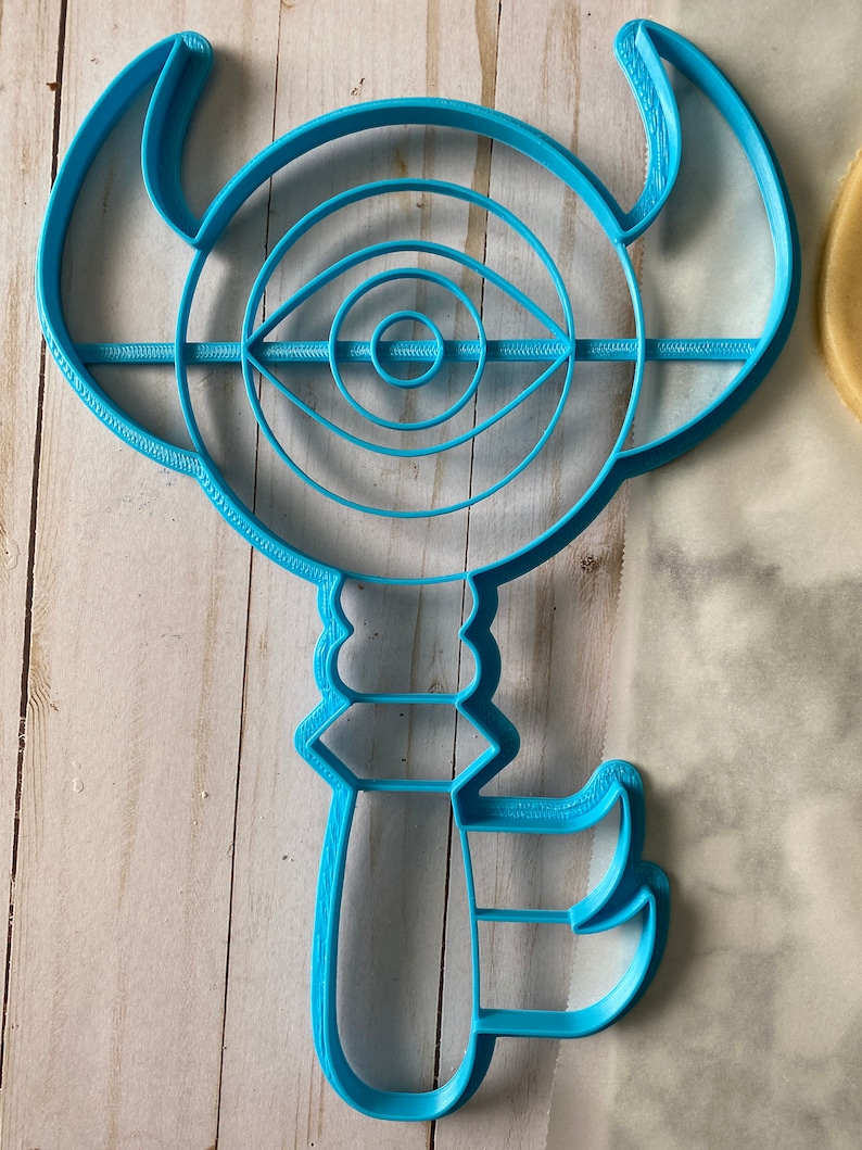 Op de afbeelding: Een blauwe plastic koekjesvormer in de vorm van een sleutel met een cirkelvormig oogontwerp. Het oog heeft concentrische cirkels en een kleine cirkel in het midden.