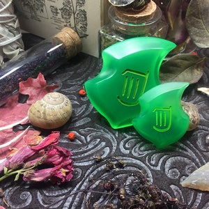 May include: Two green resin pendants with a harp symbol, one larger than the other, on a dark patterned surface with dried flowers and other natural objects.