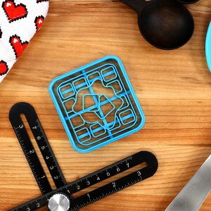 May include: A top-down shot of kitchen tools on a wooden surface. A blue cookie cutter, black measuring tools, a blue spatula, black measuring spoons, and a white oven mitt with red pixelated hearts are visible.