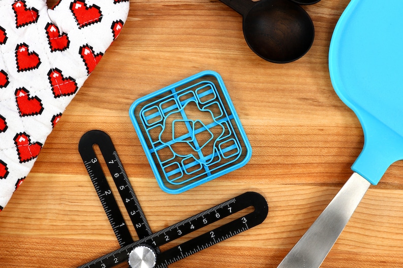 May include: A top-down shot of kitchen tools on a wooden surface. A blue cookie cutter, black measuring tools, a blue spatula, black measuring spoons, and a white oven mitt with red pixelated hearts are visible.