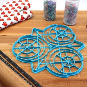 May include: A blue plastic cookie cutter shaped like three overlapping circles with a web-like design inside each circle. The cutter is on a wooden surface with a rolling pin, a ruler, and two jars of sprinkles.