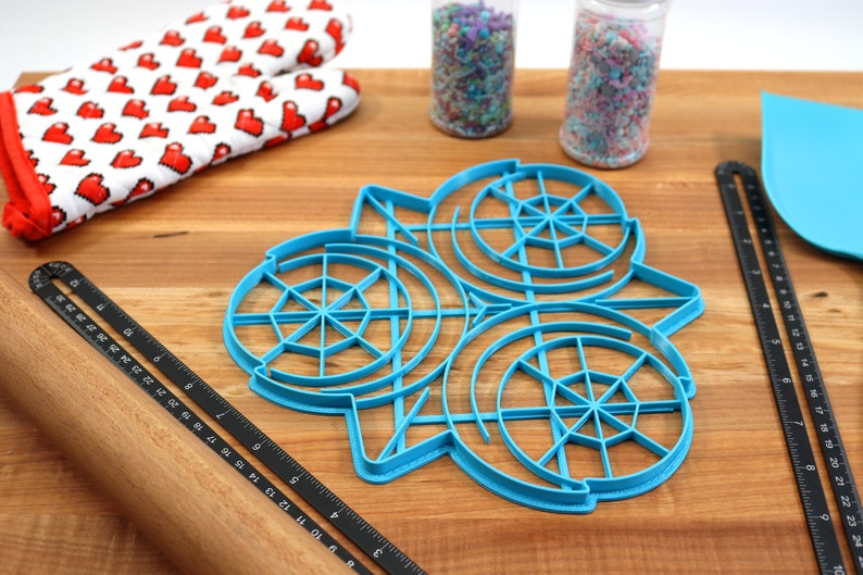 May include: A blue plastic cookie cutter shaped like three overlapping circles with a web-like design inside each circle. The cutter is on a wooden surface with a rolling pin, a ruler, and two jars of sprinkles.
