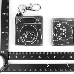 May include: Two silver metal keychains with a cartoon character design. The keychain on the left has a silver metal ring and a silver metal clasp. The keychain on the right has a silver metal clasp. Both keychains have a black and white cartoon character design with the text "X" and "SP" on the keychains.