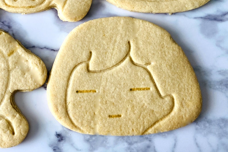 May include: A plain sugar cookie with a face cutout and yellow icing details. The cookie is on a white marble surface.