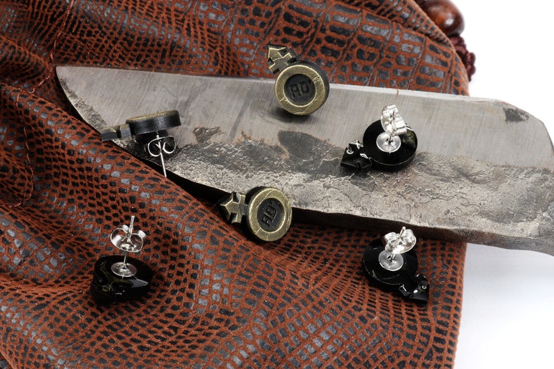 Puede incluir: Cuatro pares de pendientes con un dise&ntilde;o negro y plateado. Los pendientes est&aacute;n sobre una superficie de cuero marr&oacute;n y un cuchillo plateado.