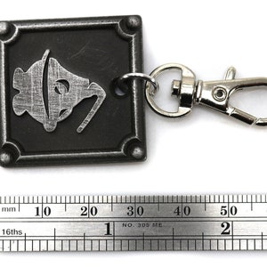 May include: A silver metal keychain with a black square charm featuring a stylized image of a bell. The charm is attached to a silver metal lobster clasp.