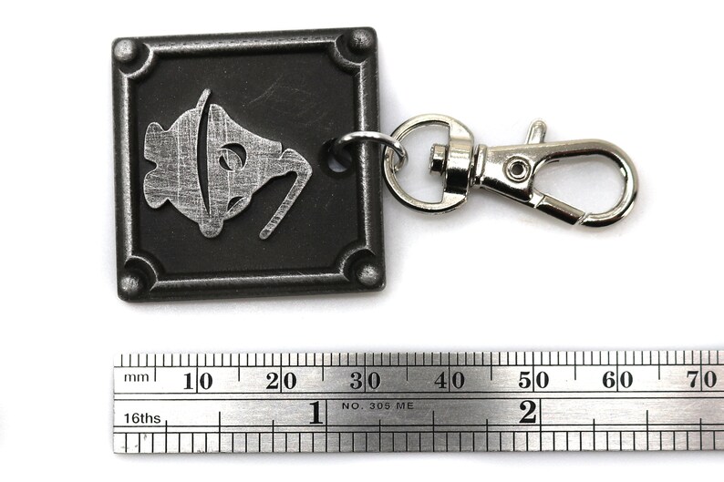 May include: A silver metal keychain with a black square charm featuring a stylized image of a bell. The charm is attached to a silver metal lobster clasp.