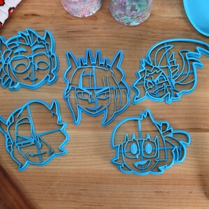 May include: Five blue plastic cookie cutters shaped like cartoon faces. The cutters are on a wooden surface.