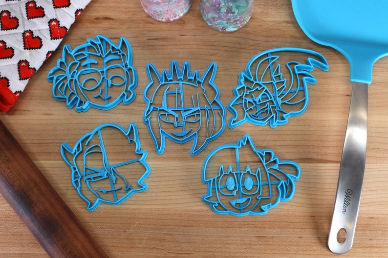 May include: Five blue plastic cookie cutters shaped like cartoon faces. The cutters are on a wooden surface.