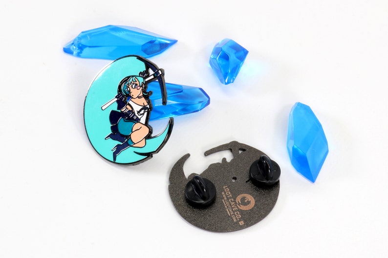 May include: A blue and black enamel pin featuring a cartoon woman with blue hair holding a pickaxe, standing on a crescent moon. The back of the pin is black with the text "Loot Cave Co." and a logo.