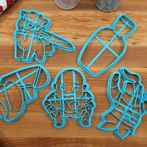 Puede incluir: Cinco cortadores de galletas de plástico azul sobre una superficie de madera. Los cortadores tienen forma de personaje de videojuego, botella, bota, persona y mano.