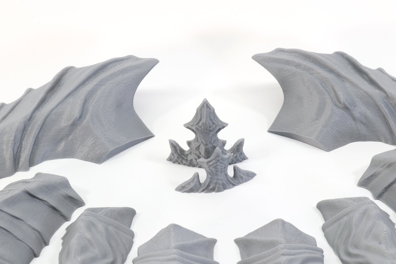 Pode incluir: Um conjunto de asas e espinhas de drag&atilde;o impressas em 3D na cor cinza. As asas s&atilde;o curvas e t&ecirc;m uma superf&iacute;cie texturizada. As espinhas s&atilde;o pontiagudas e t&ecirc;m uma borda dentada.