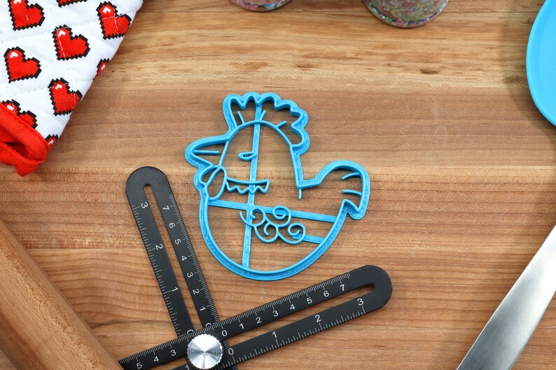 Puede incluir: Cortador de galletas de pl&aacute;stico azul con forma de pollo de dibujos animados sosteniendo un huevo. El cortador tiene un dise&ntilde;o detallado con cresta, papada y plumas.