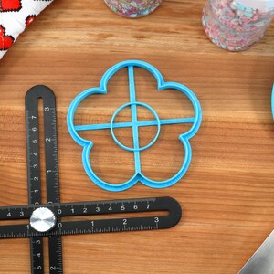 May include: A light blue flower-shaped cookie cutter, a black multi-angle measuring ruler, and a blue spatula are on a wooden surface. A white and red heart-patterned cloth and a container of sprinkles are also visible. The ruler has measurements in inches.