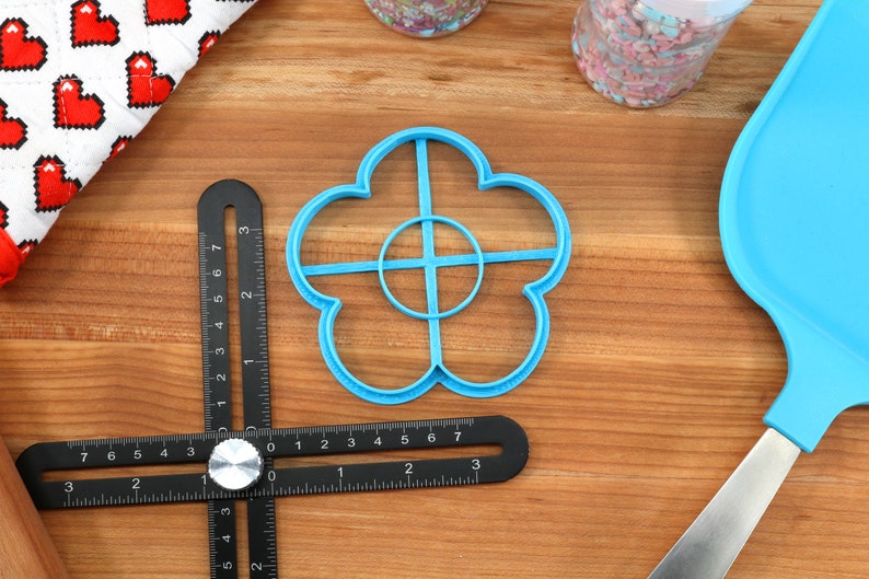 May include: A light blue flower-shaped cookie cutter, a black multi-angle measuring ruler, and a blue spatula are on a wooden surface. A white and red heart-patterned cloth and a container of sprinkles are also visible. The ruler has measurements in inches.