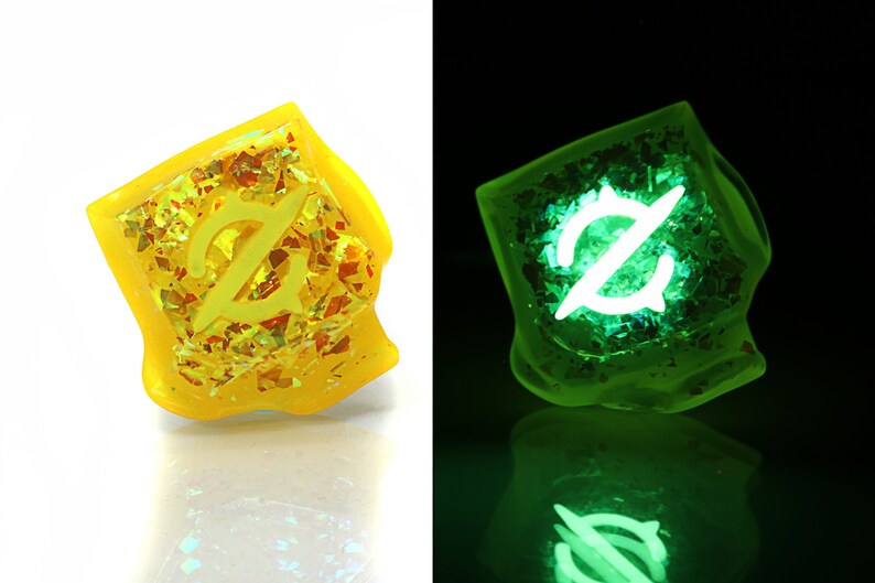 May include: A yellow dice with a white symbol on it. The dice is made of resin and has a glittery finish. The dice glows green in the dark.
