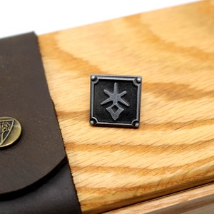 May include: A square, silver-toned metal pin with a geometric design. The pin is set against a light-colored wooden surface, with a dark brown leather strap and a brass button with a tree emblem visible in the background.