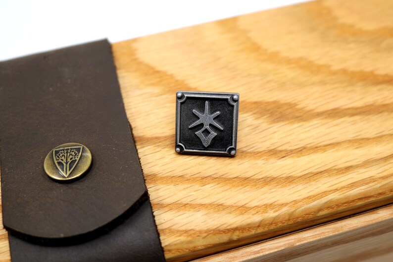 May include: A square, silver-toned metal pin with a geometric design. The pin is set against a light-colored wooden surface, with a dark brown leather strap and a brass button with a tree emblem visible in the background.