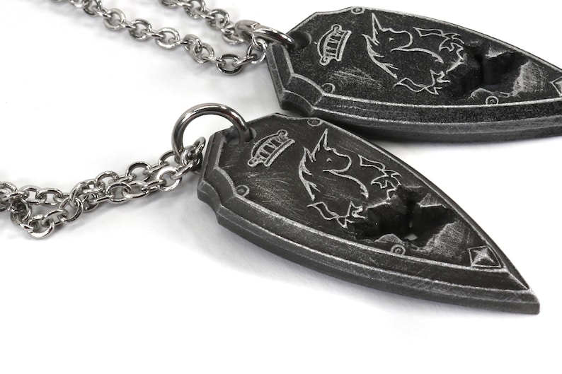 May include: Two silver necklaces with a silver chain and a pendant shaped like a shield with a unicorn design. The shield has a crown and a textured surface.