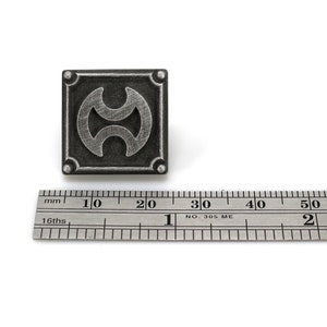 May include: A silver-colored metal pin with a black and silver design. The design is a stylized symbol with two circles and a line in the middle. The pin is about 2 cm in diameter.