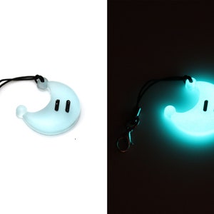 May include: A light blue glow-in-the-dark phone charm shaped like a crescent moon with two black eyes. The charm is attached to a black string with a silver clip.