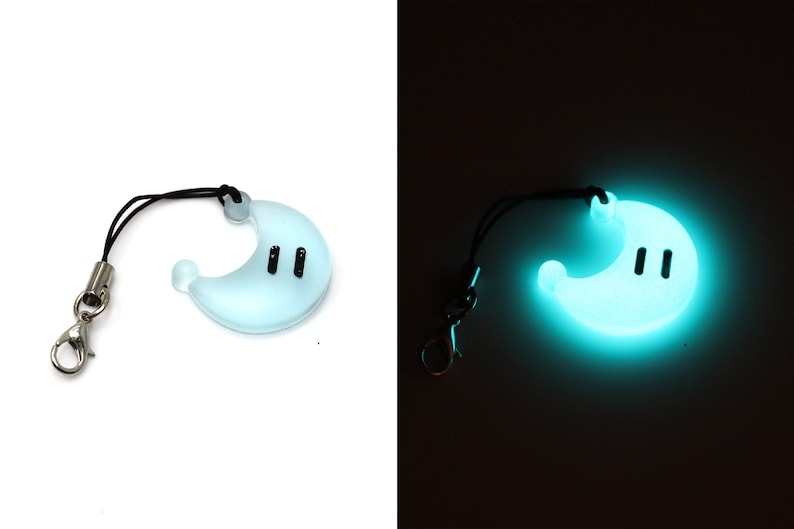 May include: A light blue glow-in-the-dark phone charm shaped like a crescent moon with two black eyes. The charm is attached to a black string with a silver clip.