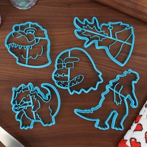 May include: Set of five blue plastic cookie cutters shaped like different dinosaurs. The cutters are on a wooden surface.