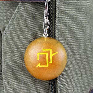 Puede incluir: Un llavero redondo de color dorado con un s&iacute;mbolo amarillo. El llavero est&aacute; unido a una cadena plateada y cuelga de una bolsa de lona verde.