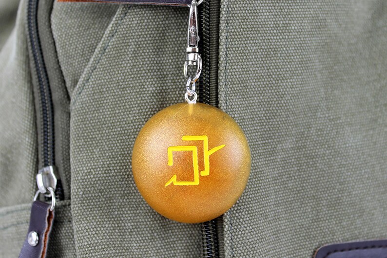 Puede incluir: Un llavero redondo de color dorado con un s&iacute;mbolo amarillo. El llavero est&aacute; unido a una cadena plateada y cuelga de una bolsa de lona verde.