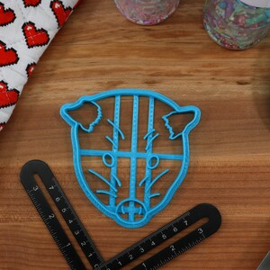 May include: Blue plastic cookie cutter shaped like an animal face with a striped pattern.