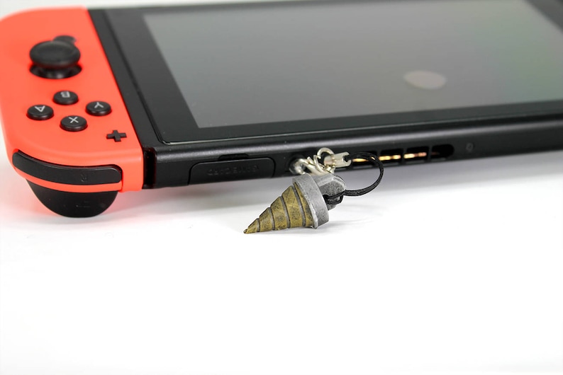 May include: A red and black Nintendo Switch console with a grey drill bit keychain attached to the console's strap.