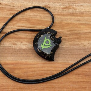 May include: Black necklace with a black and green pendant featuring a stylized eye symbol and gold flecks. The pendant is shaped like a crescent moon.
