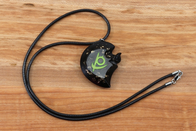 May include: Black necklace with a black and green pendant featuring a stylized eye symbol and gold flecks. The pendant is shaped like a crescent moon.