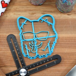 May include: A blue plastic cookie cutter shaped like a dog wearing sunglasses. The cookie cutter is on a wooden surface.