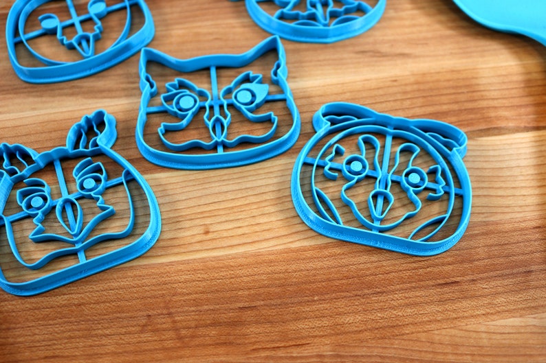 May include: Four blue plastic cookie cutters shaped like owls with different facial expressions. The cutters are on a wooden surface.