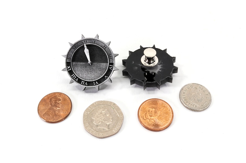 Puede incluir: Dos peque&ntilde;os relojes de metal negros en forma de engranaje con marcas de n&uacute;meros romanos. El reloj de la izquierda tiene una aguja blanca que apunta al n&uacute;mero XII. El reloj de la derecha tiene un pasador plateado en el centro. Cuatro monedas est&aacute;n dispuestas debajo de los relojes.