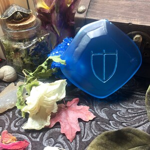 May include: A blue, faceted, glass shield with a white, stylized sword and shield design. The shield is on a dark, patterned surface with dried flowers and leaves.