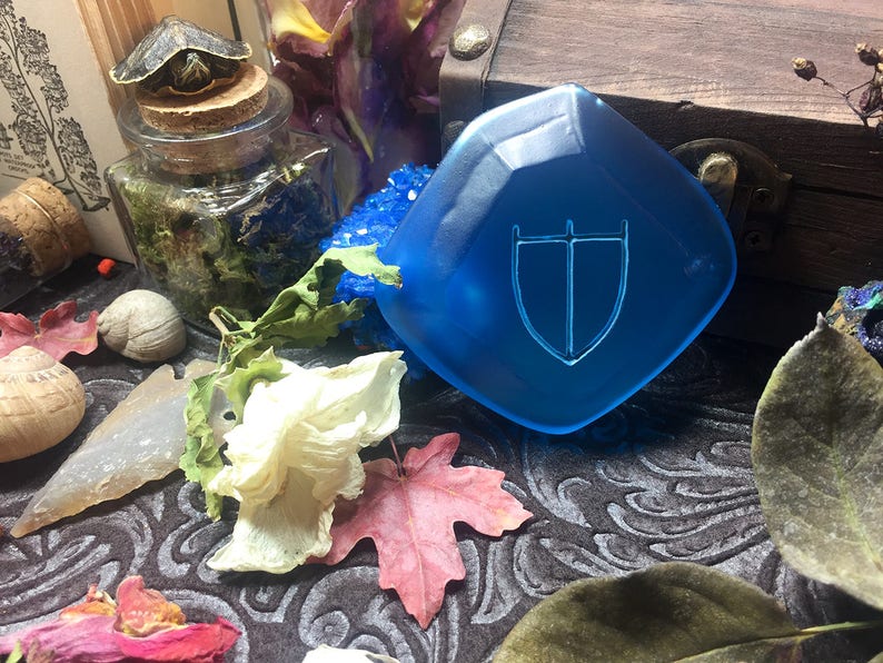 May include: A blue, faceted, glass shield with a white, stylized sword and shield design. The shield is on a dark, patterned surface with dried flowers and leaves.